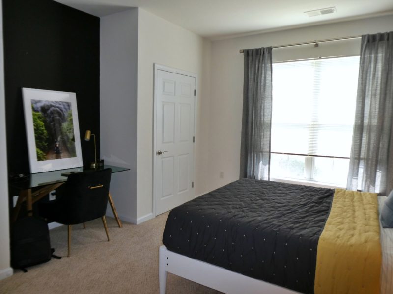 This image shows the bedroom area showcasing the dark tone wall color, elegant bedding, accessible working station, and an overlooking view outside the apartment.